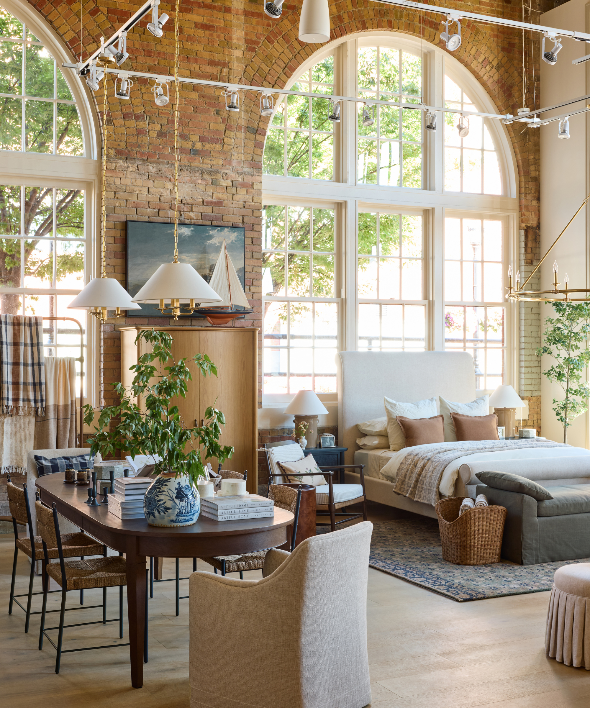 Charming brick-walled interior shot of the new McGee &amp;amp; Co. flagship, featuring a cozy bedroom set up alongside a dining table vignette with vintage-inspired upholstered and woven furniture.
