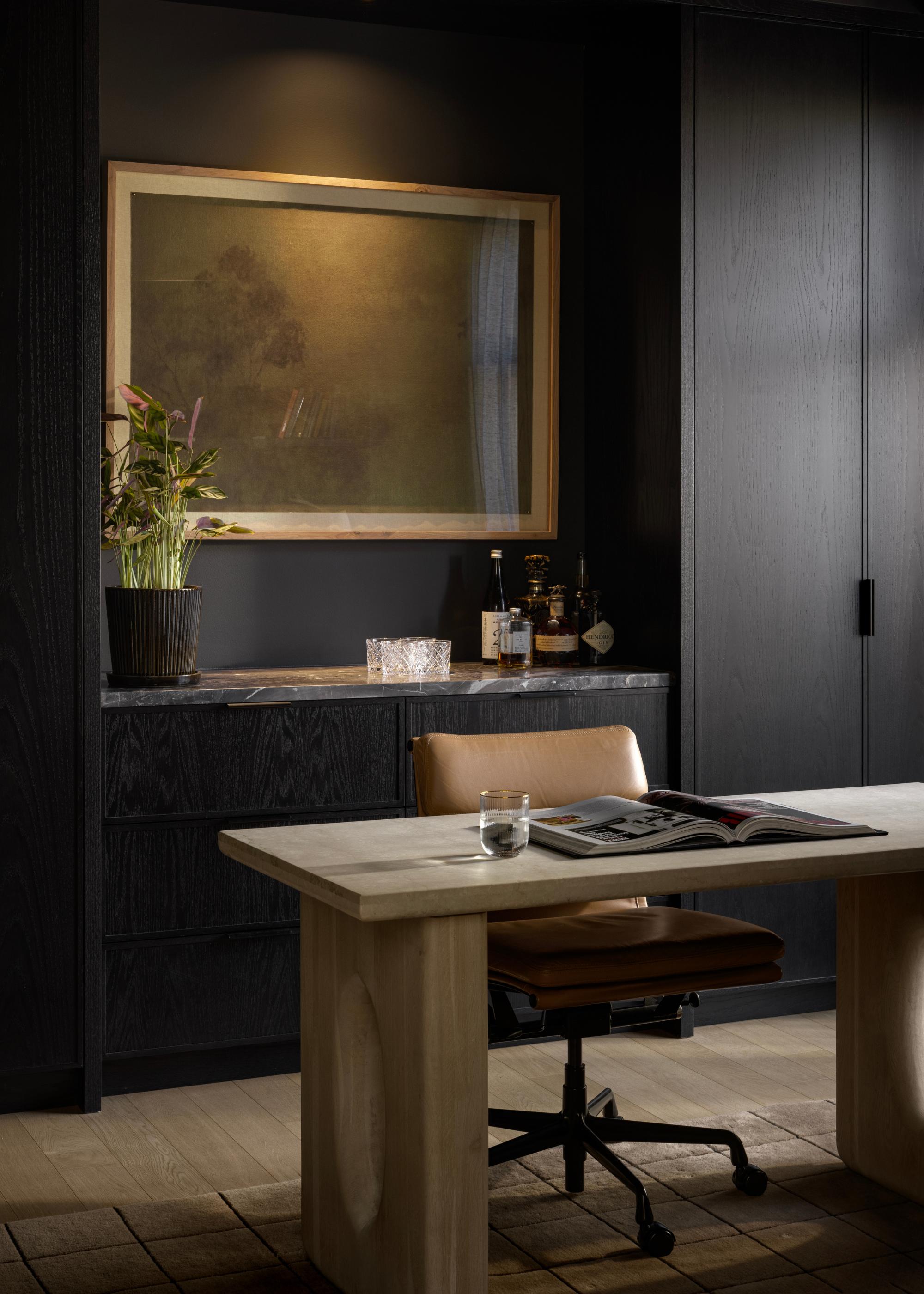 A moody home office with a potted plant, a framed painting, a leather swivel chair, a stone desk, and an open book