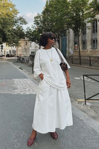 a photo showing what shoes to wear with skirts with a woman standing in the street wearing a white t-shirt with a white midi skirt and brown ballet flats