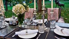 Outdoor dining table with silver flatware, candelabras and napkin rings. There are peonies in vases and pink upholstered chairs in view