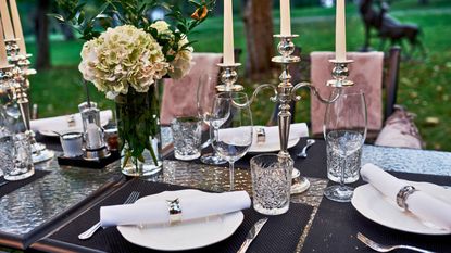 Outdoor dining table with silver flatware, candelabras and napkin rings. There are peonies in vases and pink upholstered chairs in view