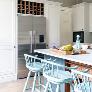 Bank of tall units with a stainless steel fridge freezer behind a kitchen island with blue wooden bar stools