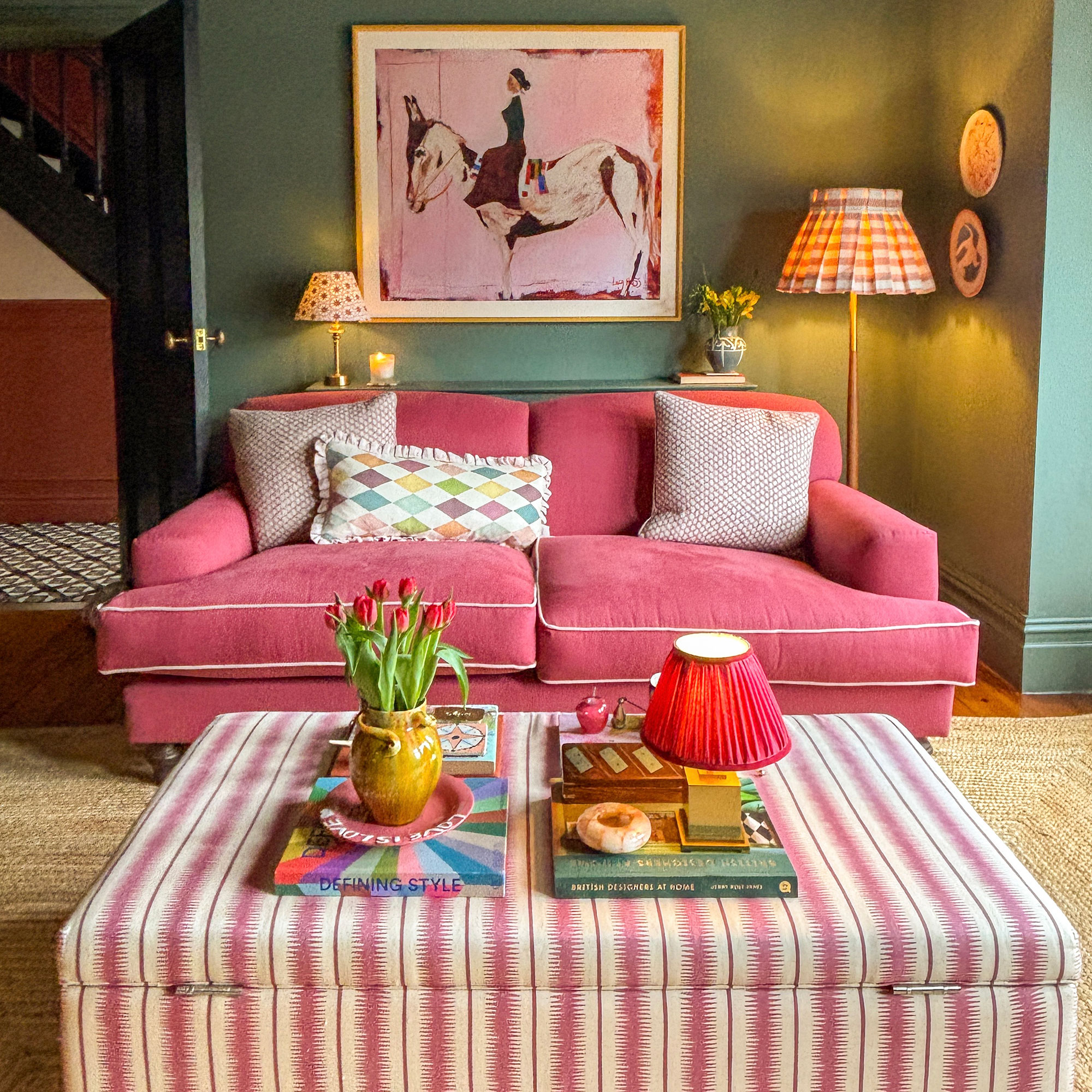 Green living room with pink sofa, pink painting with horse and pink and white striped fabric ottoman acting as coffee table