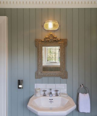 small bathroom with pale blue wall panelling and ornate mirror with industrial style light above