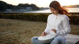 Roland Mood Pan lifestyle shot: a young woman with dark hair wearing a white shirt and blue jeans is tapping a Mood Pan while sitting on grass. A bay is behind her and the sun is going down.