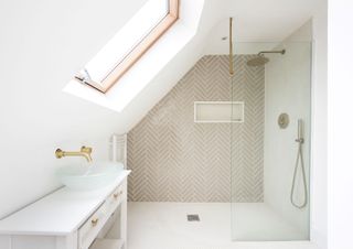 An ensuite bathroom in a loft conversion with a sloped ceiling to the left of the picture with a sink under it and a shower to the rear with beige tiling on the far wall