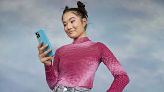 Feminine-presenting Gen Z person wearing a dotted pink top, with pink eyeshadow and space buns, holding a blue smartphone