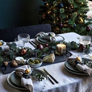 close up of a dining table beside a christmas tree with soft napkins decorated with baubles and a large cheese board centerpiece