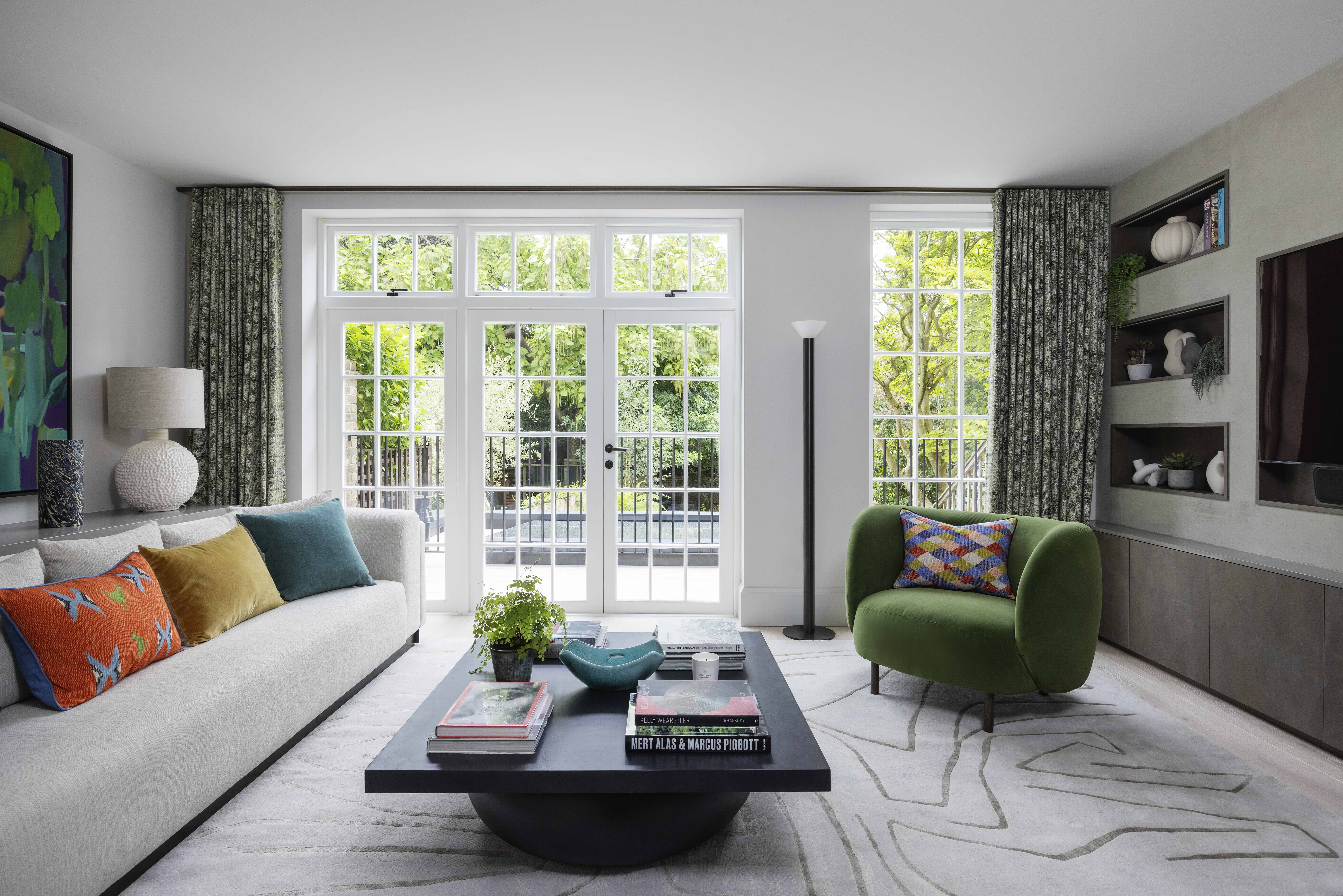 modern living room with white walls, french doors, patterned curtains on either side, a built-in media unit, green armchair, large black coffee table styled with books and a potted plant, patterned gray rug, and large gray sofa with colorful cushions