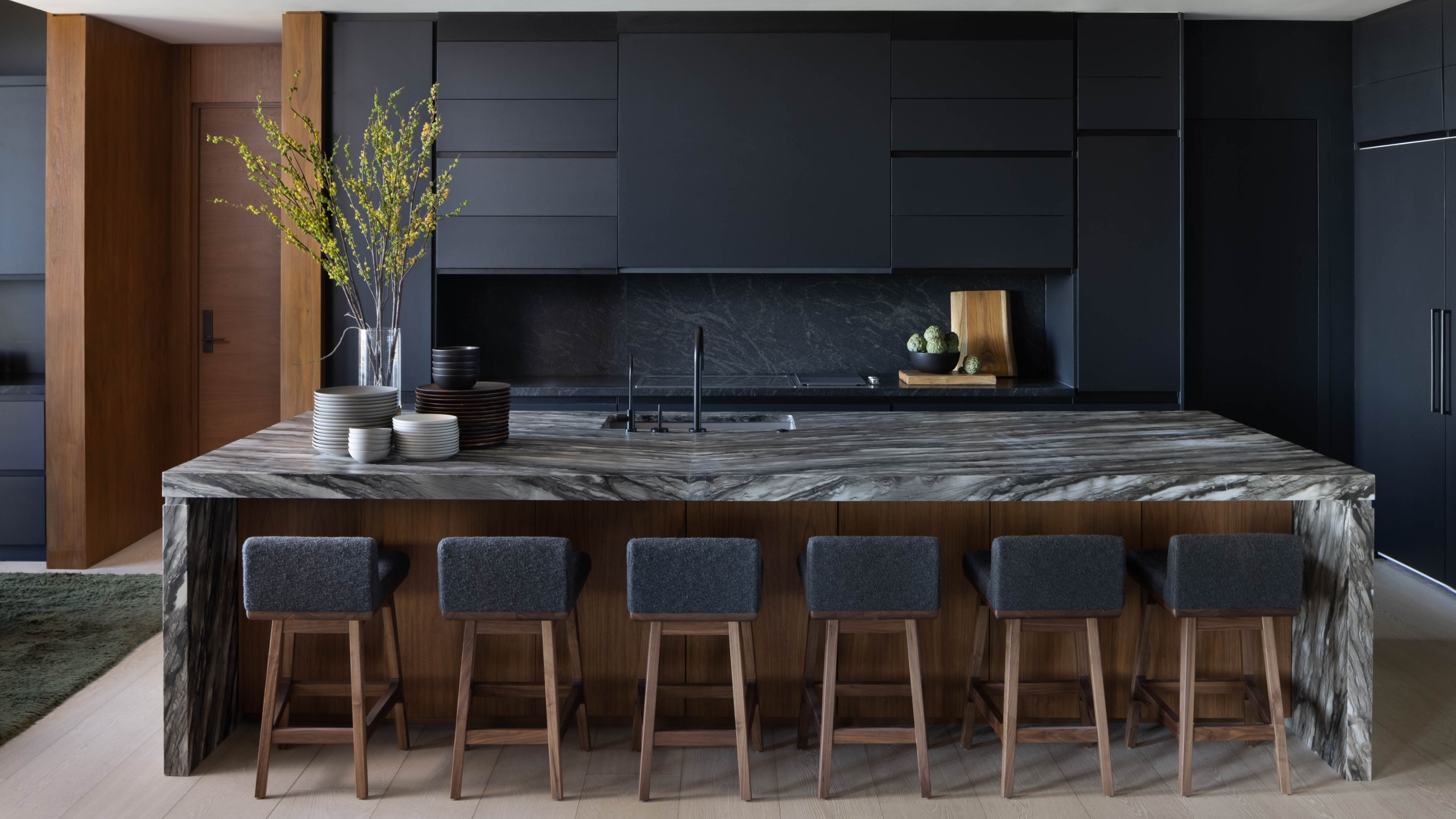 A modern dark off-black kitchen with an oversized marble waterfall island and six bar stools line up against it