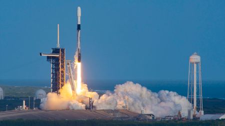 a white and black rocket launches into a clear blue sky from its ocean side launch pad