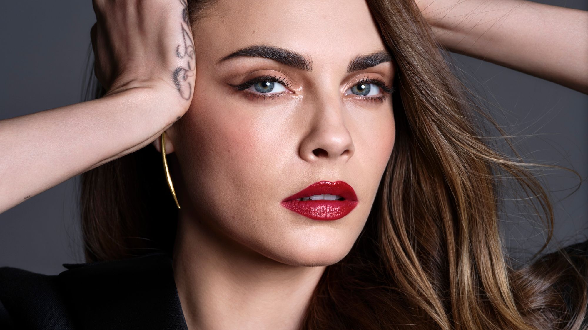 An image of Cara Delevingne wearing red lipstick and winged eyeliner against a gray backdrop.