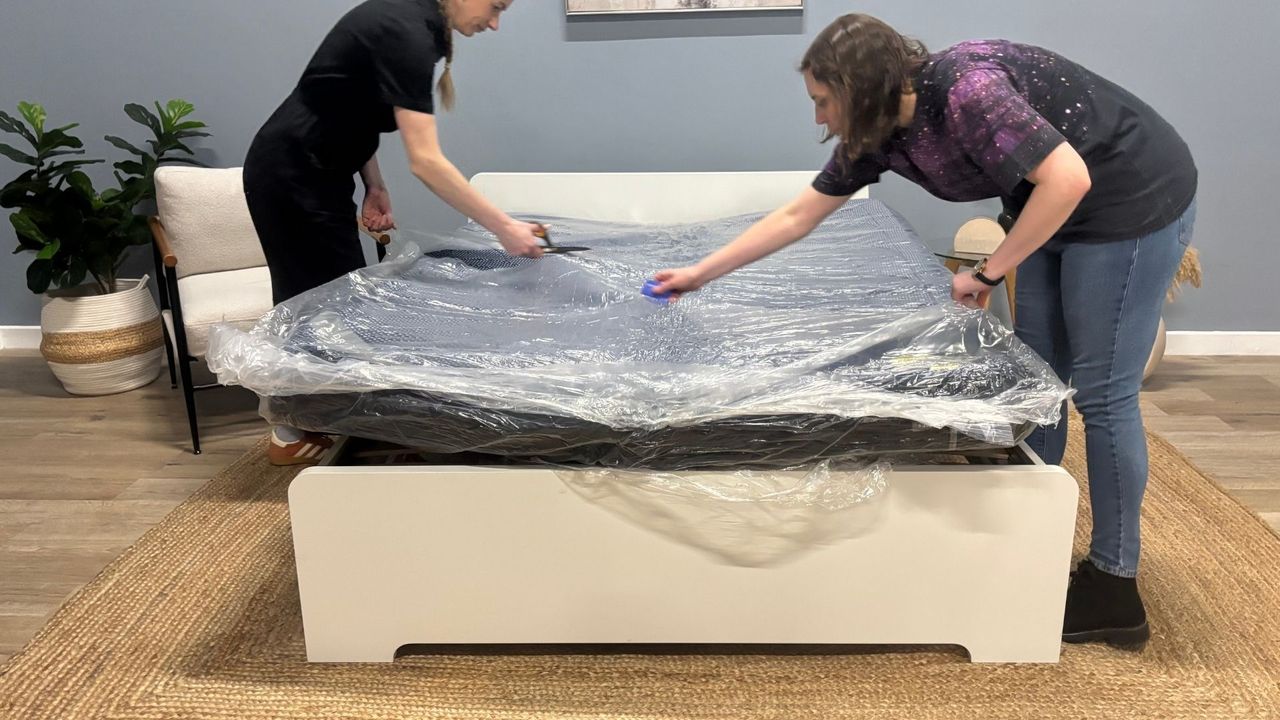 Two mattress testes unbox the DreamCloud Luxe Memory Foam mattress, taking it out of its box, cutting it out of its plastic wrapping and placing it on a white bed frame