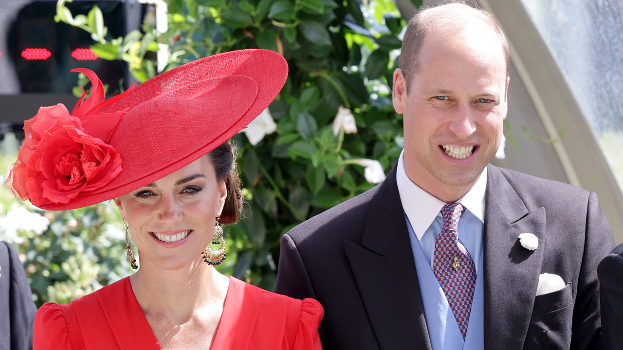 Kate Middleton wears a red hat with a wide brim and a matching red wrap dress, while Prince William wears a suit and waistcoat