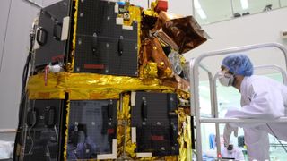 a person in a white coat, a mask and blue hairnet inspects a large black-and-gold spacecraft inside a white clean room