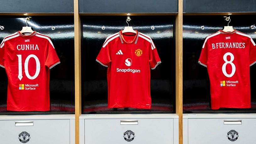 Three red Manchester United soccer jerseys hanging in a locker room, displaying front Snapdragon branding and back Microsoft Surface logos below player names