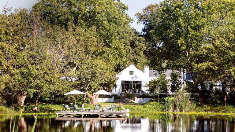exterior of white farmhouse in South Africa surrounded by oak trees with a lake and with outdoor seating area on a jetty with parasols