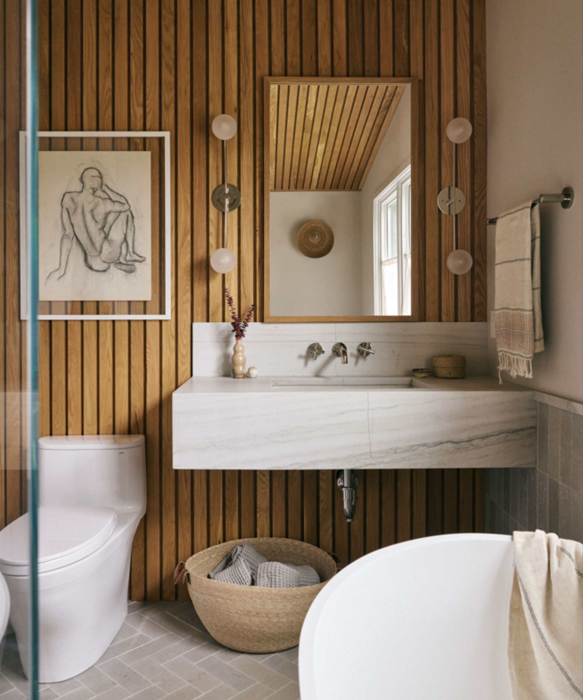 small bathroom with wooden pabelled walls, marble sinks, mirror, wall lights and artwork on wall, basket under sink with towels, corner of bath in lower right hand side