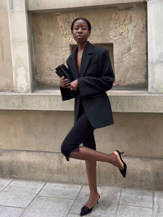 French fashion influencer Sylvie Mus in Paris wearing an oversize black blazer, black capri pants, and black Manolo Blahnik mules and carrying a black clutch.