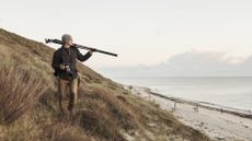 Man walking along sand dunes, looking out to sea, with one of the best travel tripods on his shoulder