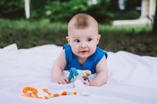 Close up photo of smiling dark-eyed baby boy in blue t-shirt lying on stomach with his doll and toy on white sheet outdoors in summer day.