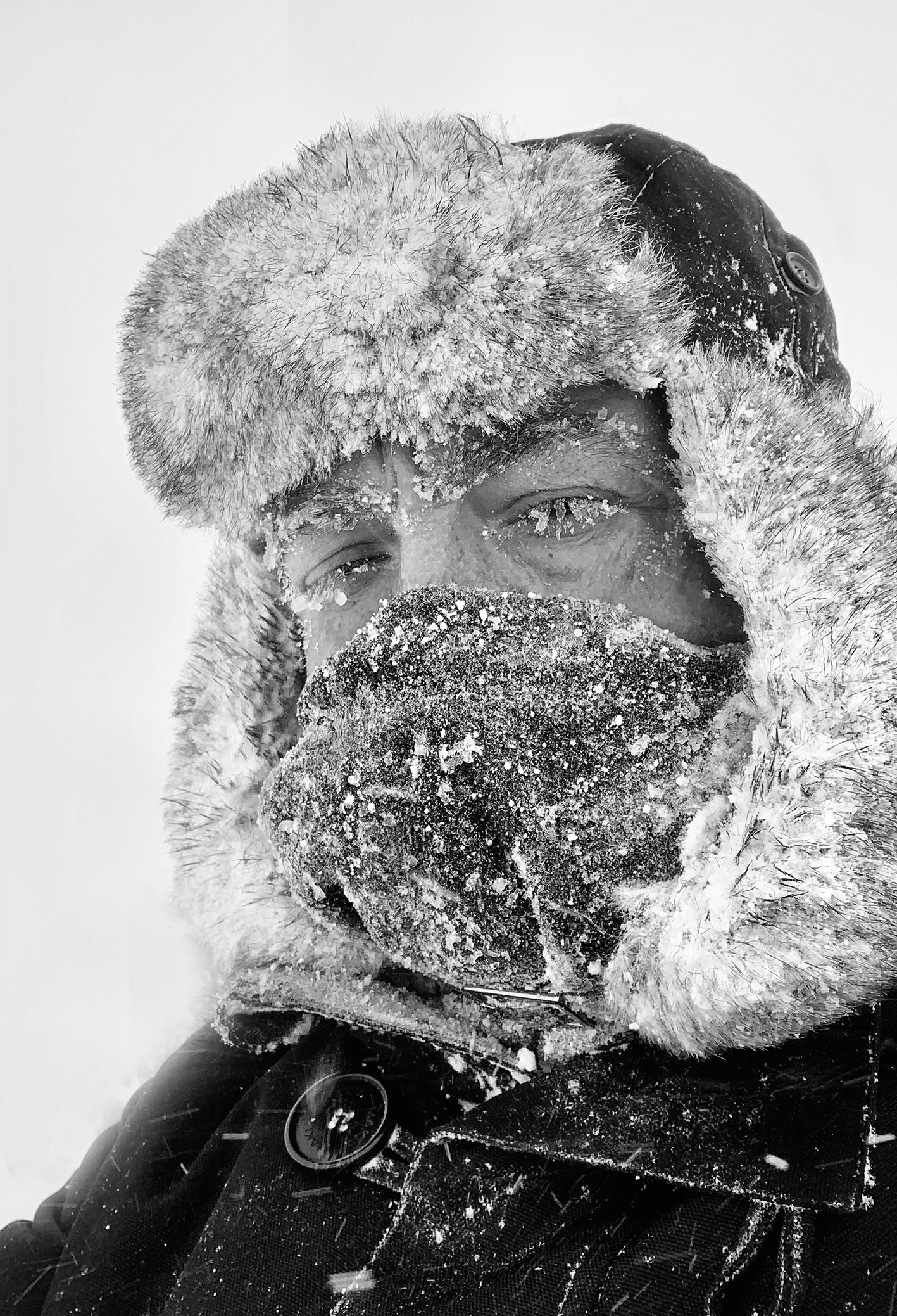 Photographer David Gibbon, wearing a fur-lined winter hat and snow-covered jacket, looking up in a snowy environment, black and white photo