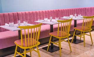 Interior view of the dining area at The Magnolia Hotel featuring a blue wall, row of pink seats, light coloured square tables with tableware, yellow chairs and tiled floors