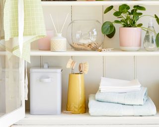 a white cupboard full of plants, containers, towels and linens, and a candle and a diffuser, with a glass door