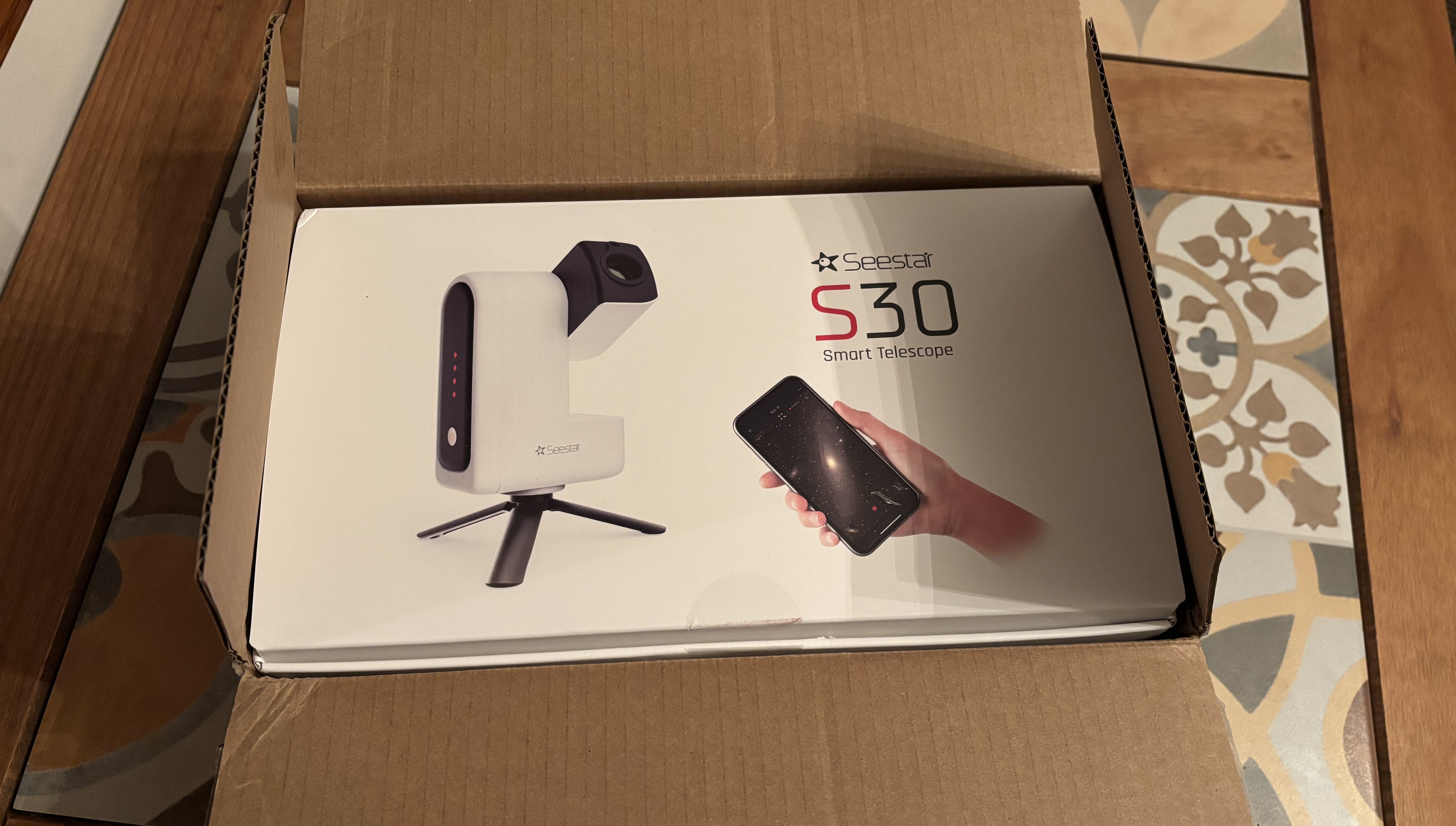 A white cover featuring the ZWO Seestar S30 smart telescope and a smartphone is inside an opened cardboard box.