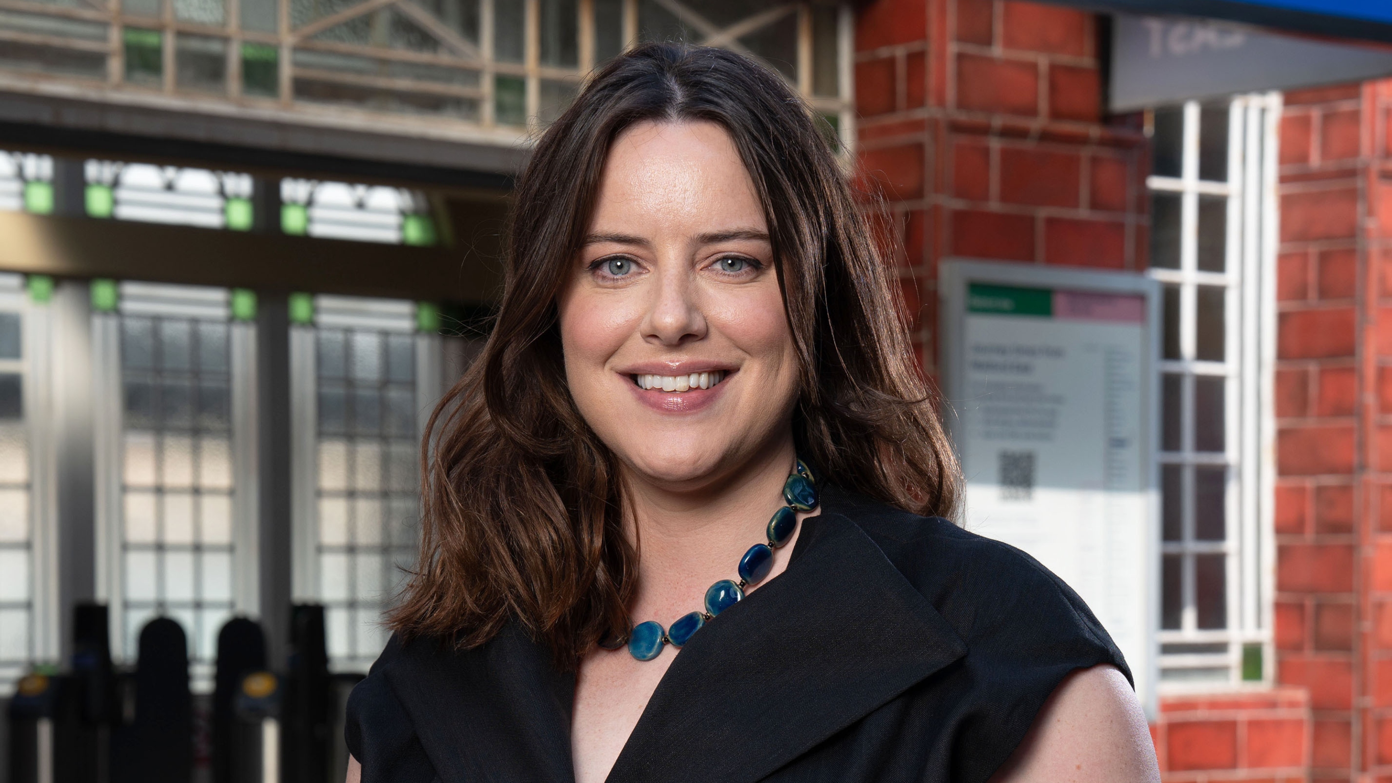 Michelle Ryan as Zoe Slater standing in front of Walford tube station and smiling, wearing a black top and blue bead necklace