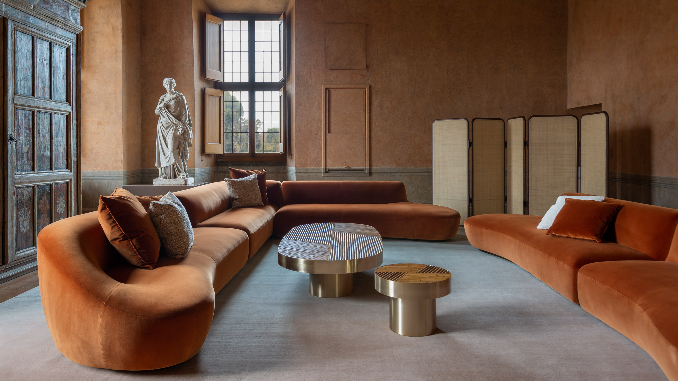 An orange-tinted historical reception room furnished with contemporary, sculptural designs, including a chrome-clad coffee table, velvety oversized couches, and room dividers.