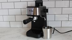 Mr. Coffee Steam Espresso Maker with stainless steel metal milk frothing pitcher on marble kitchen countertop with white tiled kitchen wall in background