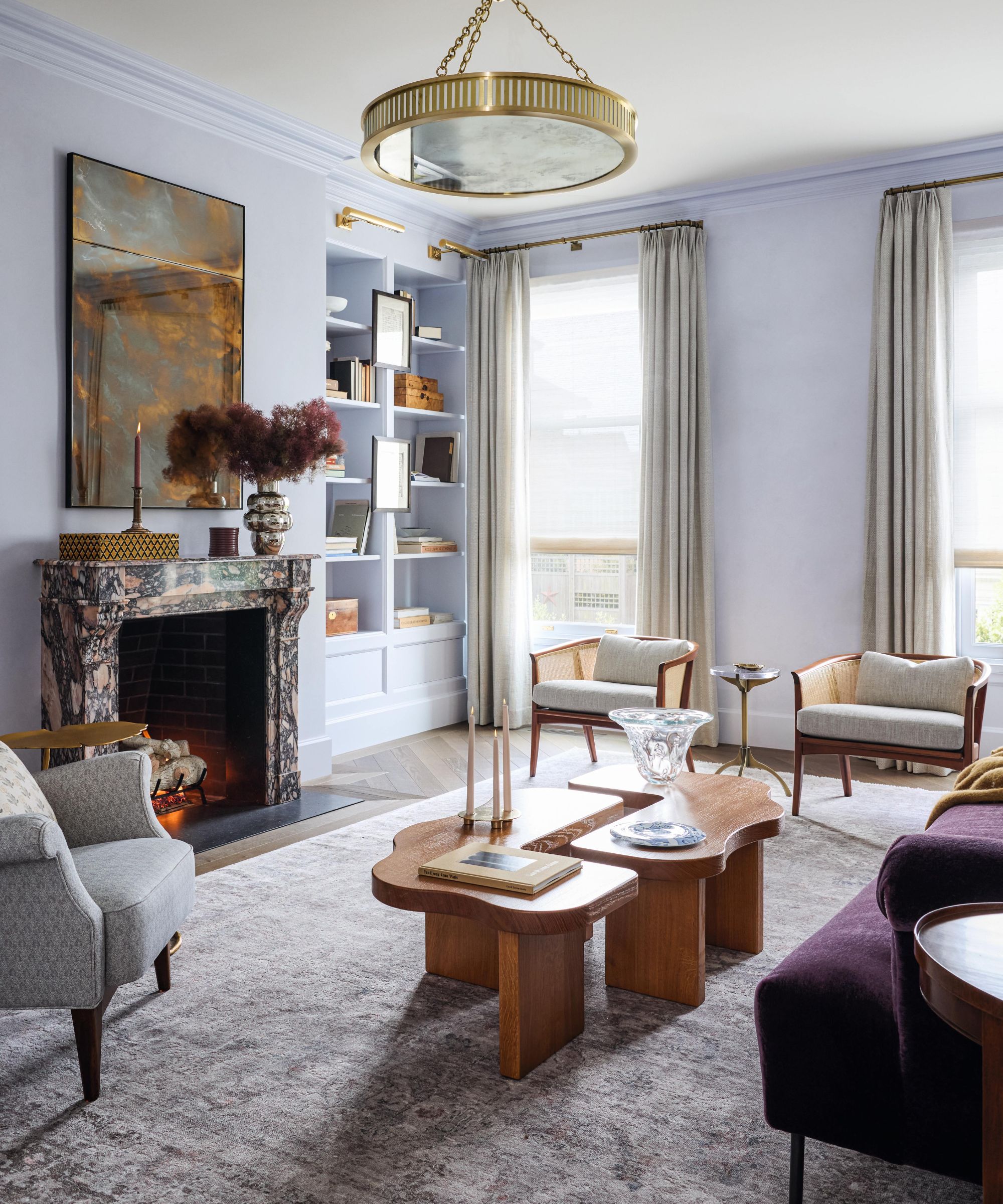 a light blue edwardian living room with two large windows, marble fireplace, purple sofa, curved wooden coffee tables and eclectic chairs