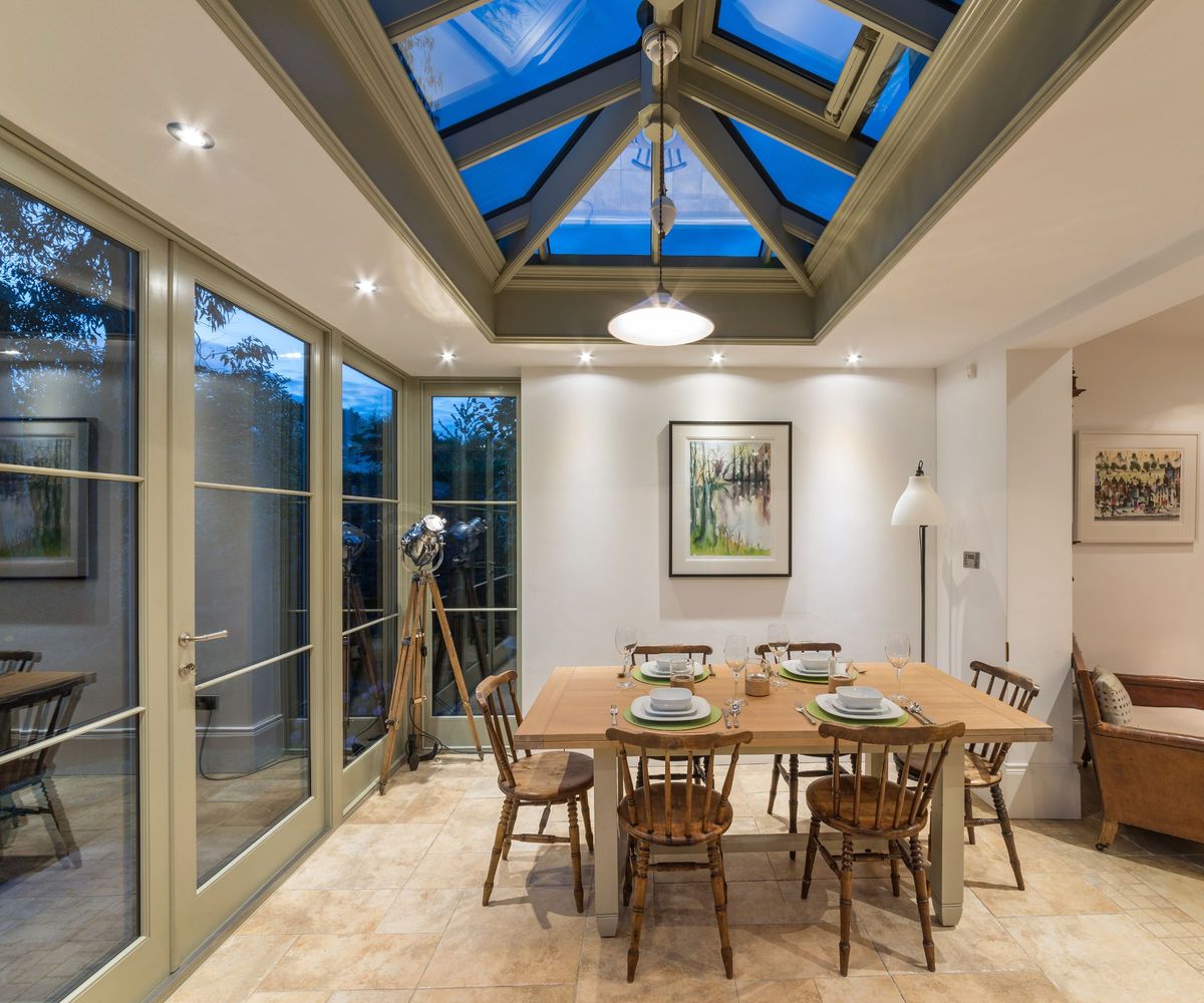 Inside the orangery at dusk, a farmhouse table with Windsor chairs sits beneath a dramatic lit glass lantern roof, with framed watercolours and a tripod floor lamp against white walls.