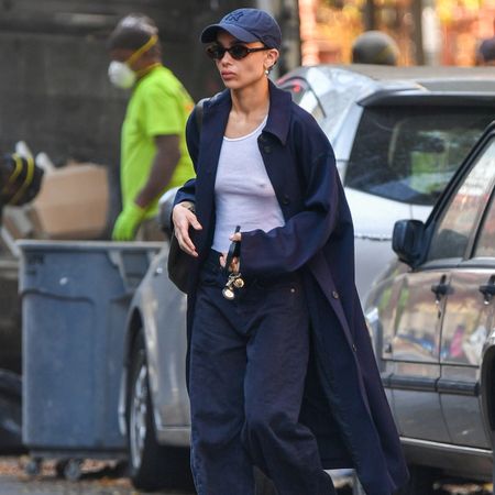 Zoe Kravitz in NYC wearing a navy baseball cap, sunglasses, a white tank top, blue long coat, navy jeans, and the Row Zipped boots in black.