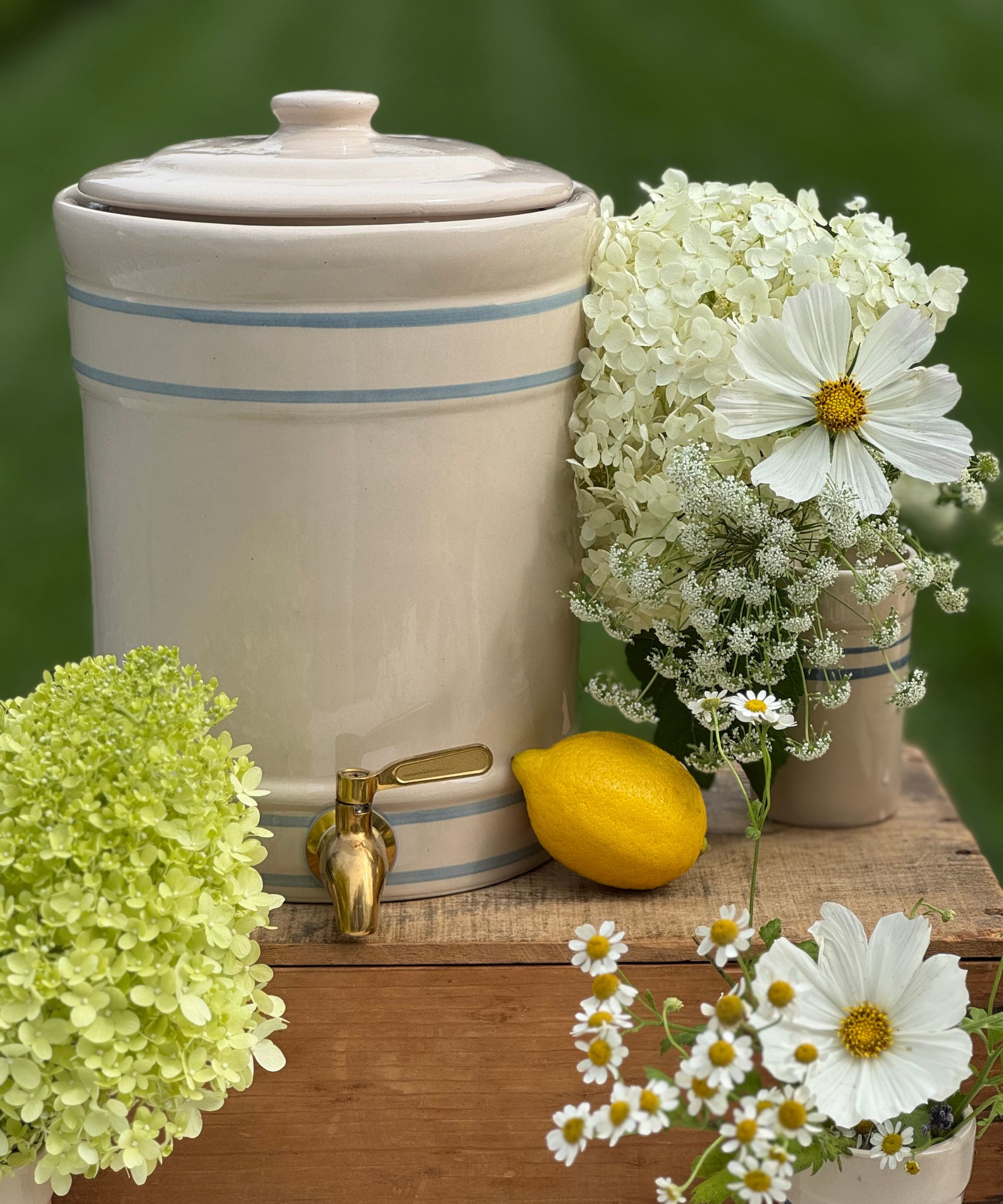 Ceramic drinks dispenser on a wooden crate with vases of flowers and a lemon