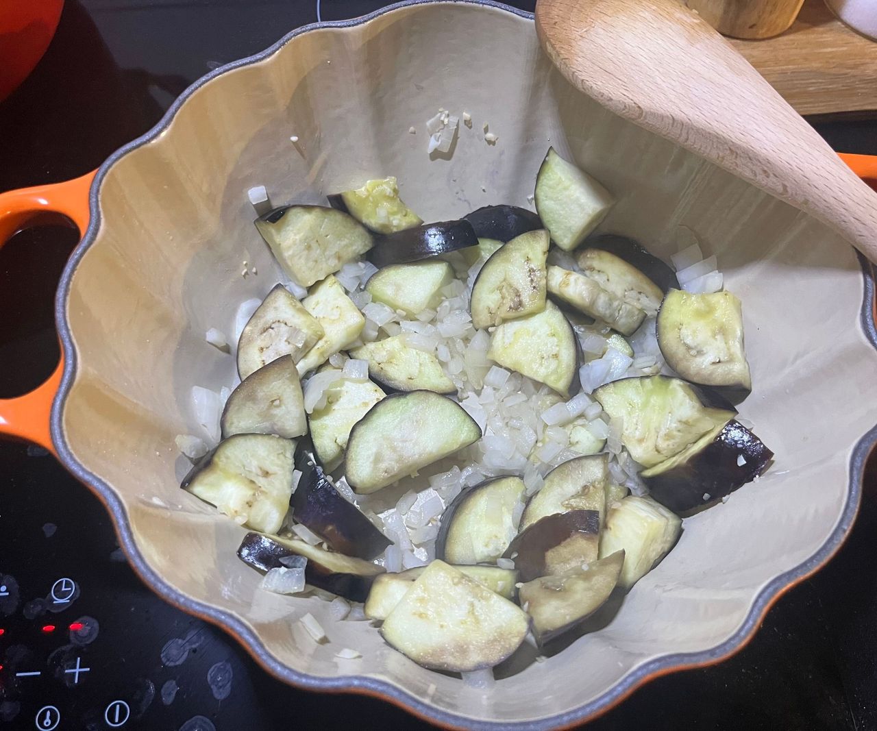 An orange Le Creuset Pumpkin cocotte on a black stove with onions and aubergine chunks inside.
