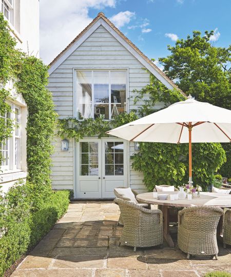 Exterior of a shiplap house with a patio that features a wicker outdoor furniture set, a wooden dining table, a cream parasol, and a tiled floor, with hedges and box planting framing the area.