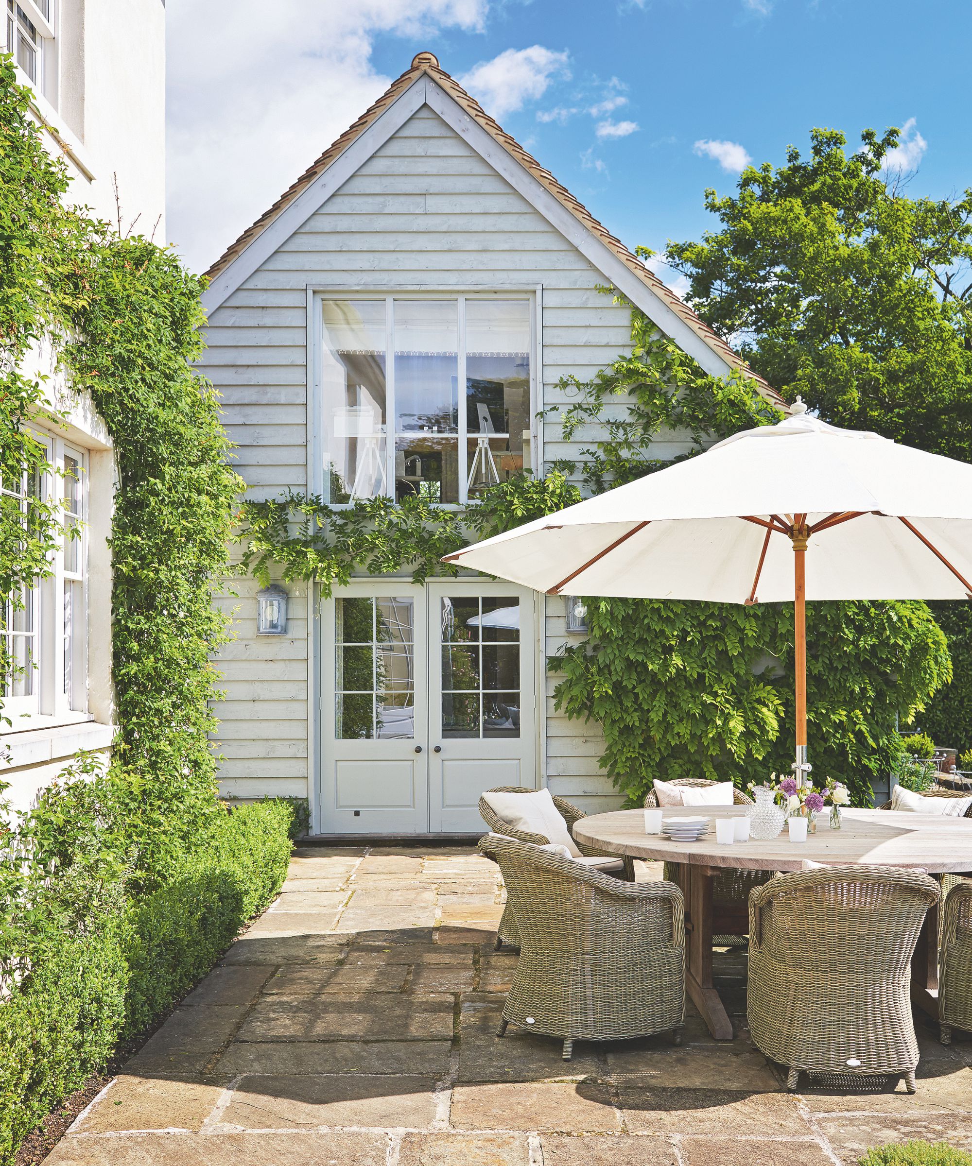 Exterior of a shiplap house with a patio that features a wicker outdoor furniture set, a wooden dining table, a cream parasol, and a tiled floor, with hedges and box planting framing the area.
