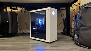 The HP OMEN 25L gaming desktop PC in white, sitting on a wooden floor with its glass panel facing forward and its RGB-light fan turned on.