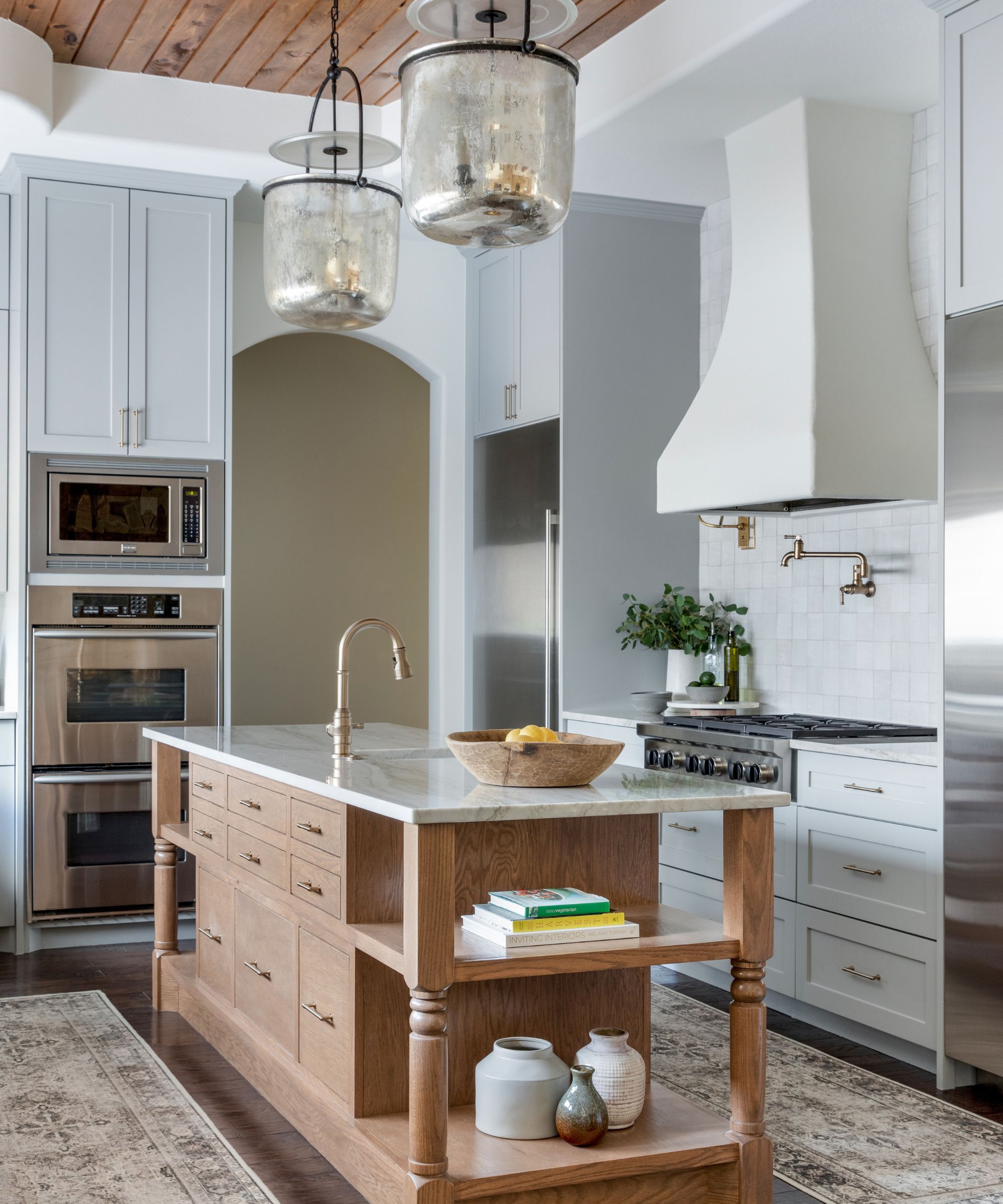 A large modern kitchen with pale blue cabinets, a wooden island with a white countertop, stainless steel appliances, brass fixtures, glass pendant lights and two neutral vintage-inspired runner rugs.