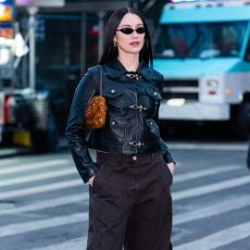 Lola Tung stands on the road in New York wearing a leather toggle jacket with low-rise jeans, cow print trainers and a furry bag.