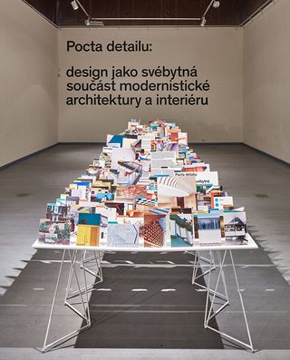 Front view of the show at the Prague Academy of Arts and Design featuring a long table filled with upright photographs in a room with white walls and grey flooring. There is black text on the back wall