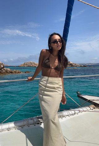 a photo showing what to wear on a boat with a woman wearing brown sunglasses, gold hoops, a brown bikini, and a column tan linen maxi skirt