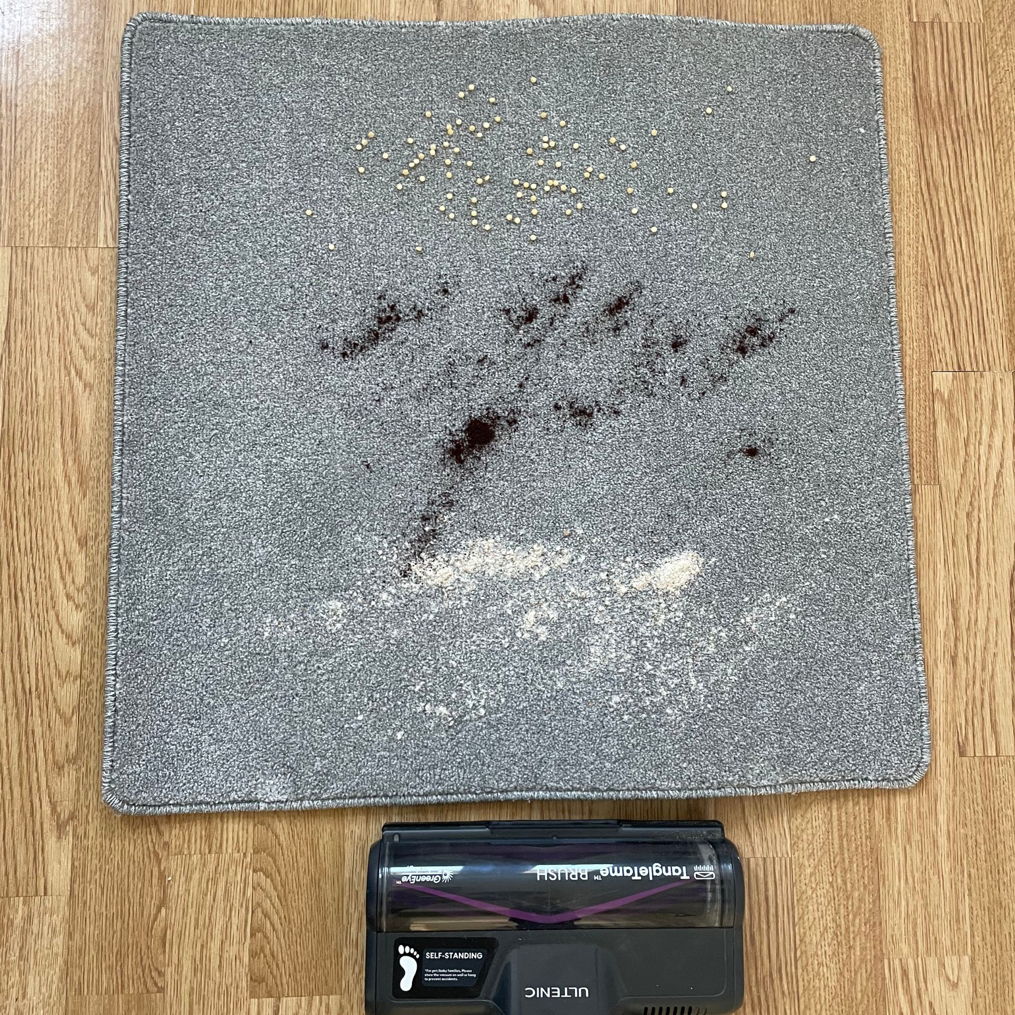 Ultenic U20 cordless vacuum behind a carpet sample covered in cous cous, coffee grounds, and flour