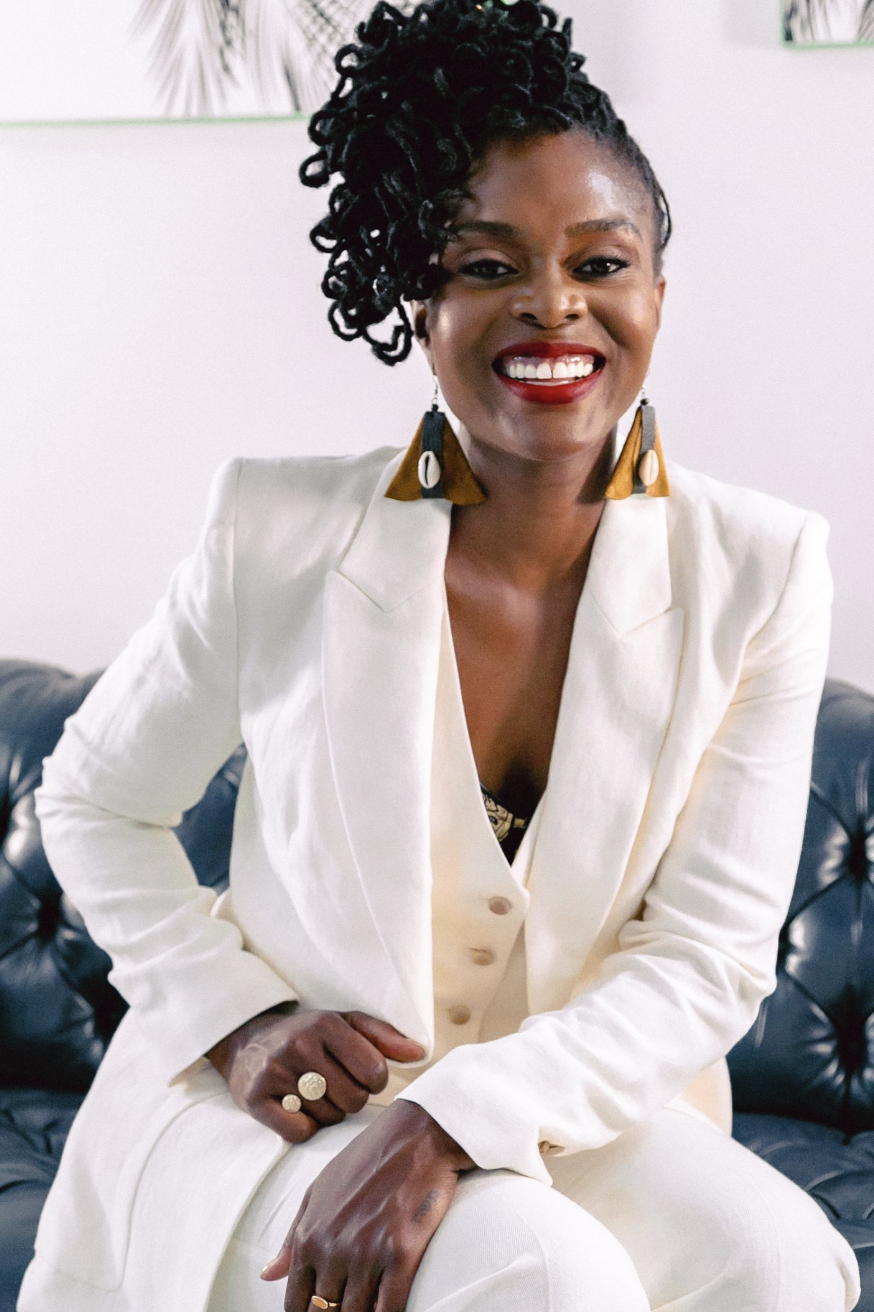 a headshot of a black woman with locs wearing a white pantsuit with her hair styled in a high updo
