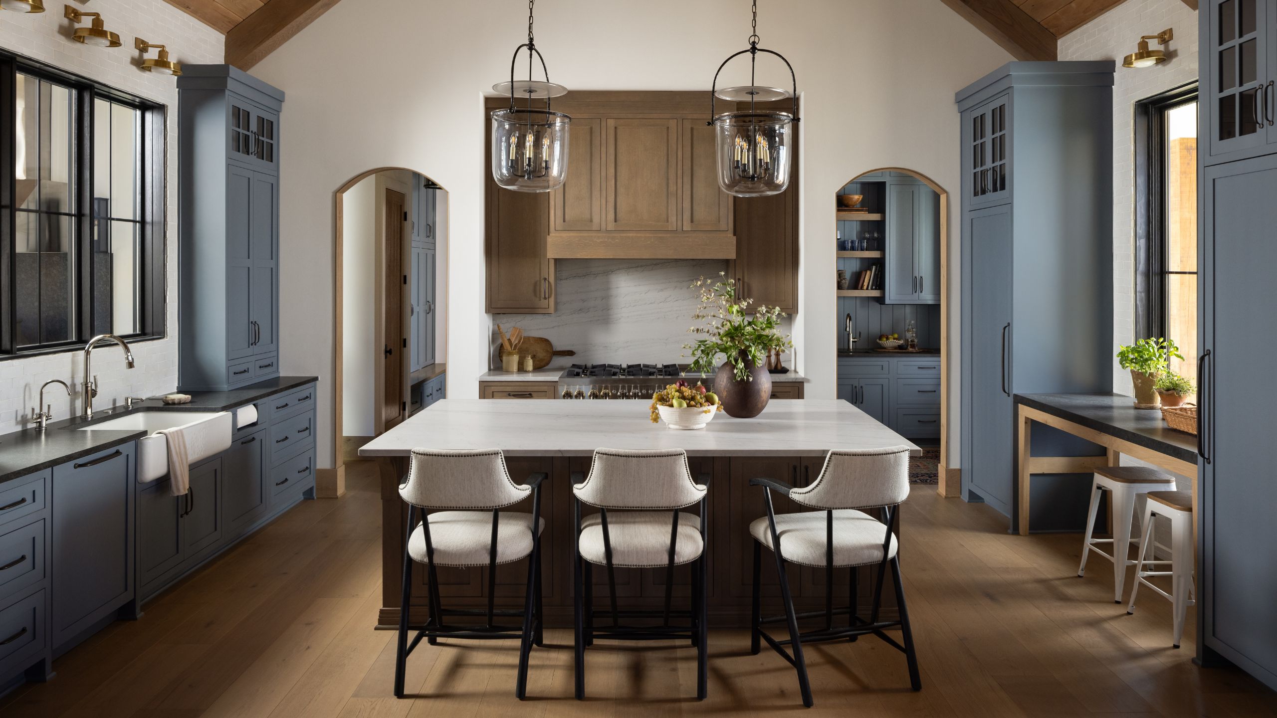 Spacious rustic-modern kitchen with vaulted wood ceiling, blue cabinets, marble island with three white counter stools, and glass pendant lights.