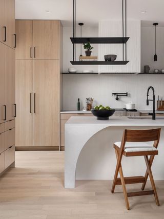 kitchen with wooden cabinets with pale wood floor and white walls and white island with curved edge and black hanging shelf and wooden stool