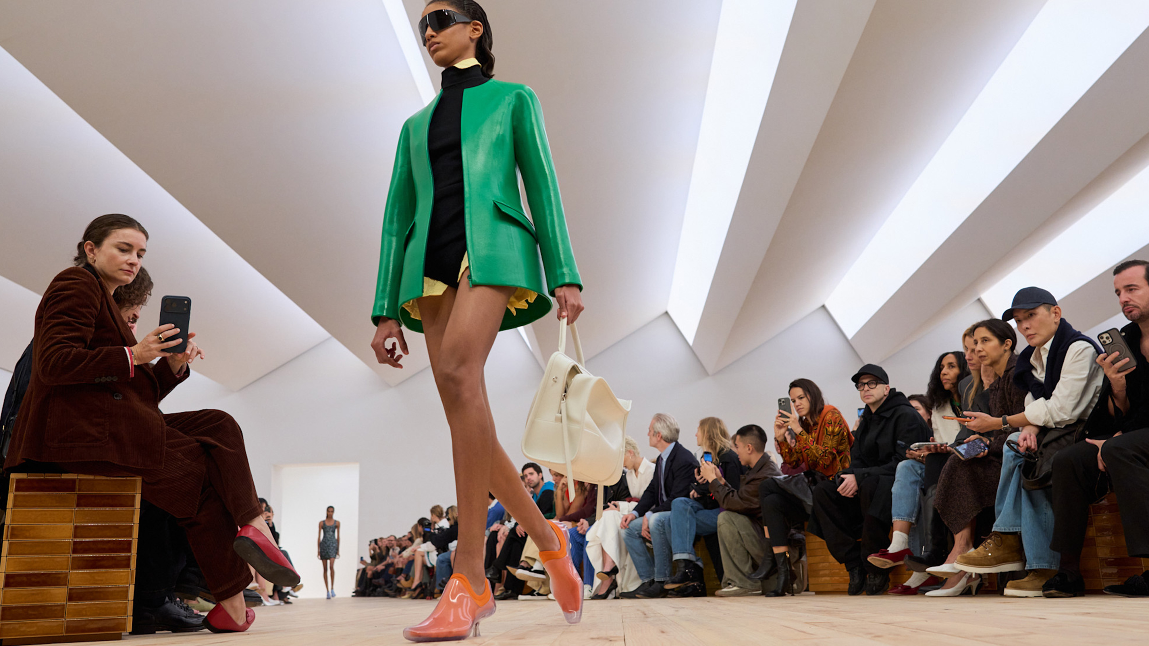 A model at the SS26 Loewe show wearing a green leather sculpted blazer with a sweater and white bag.