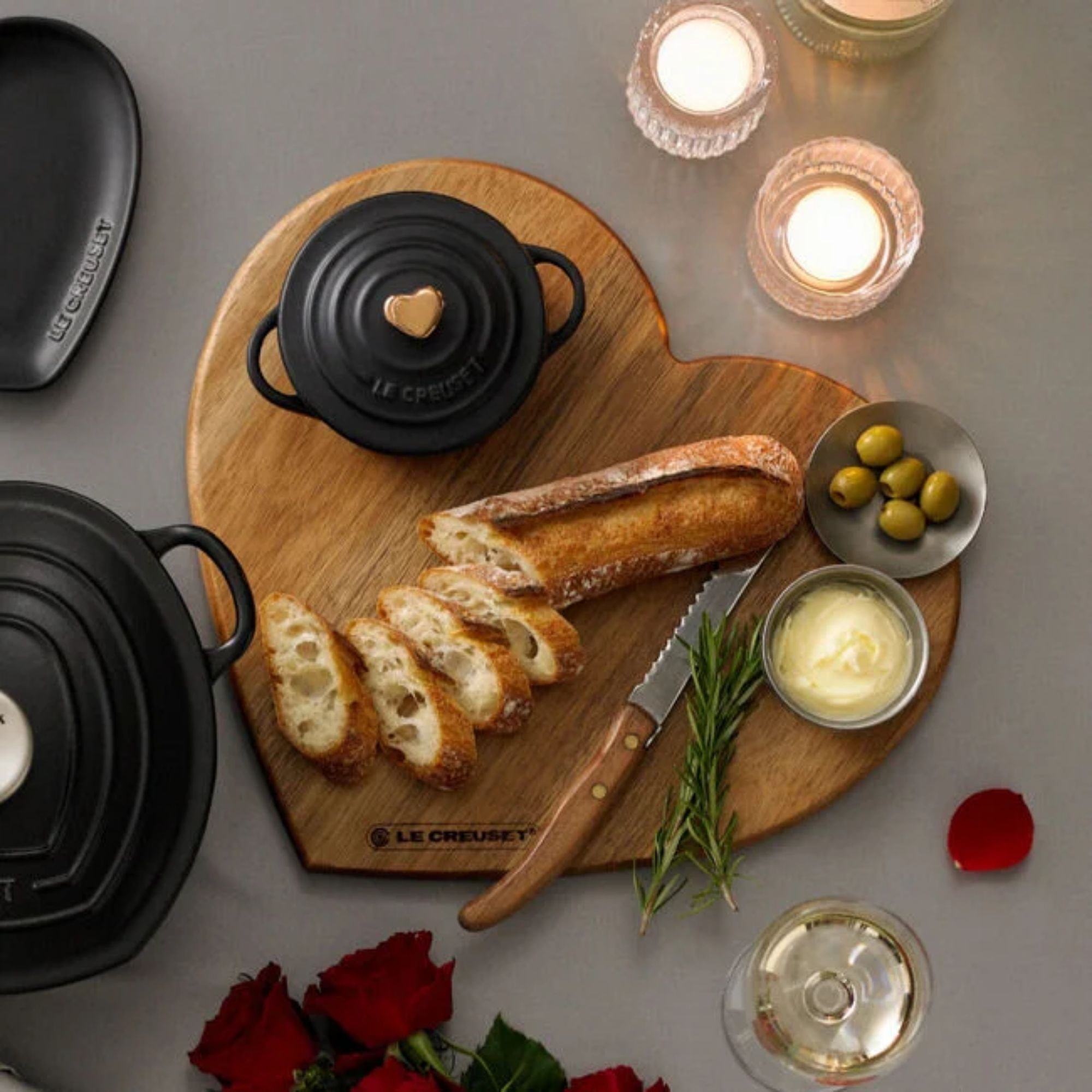 A heart shaped wooden cutting board with a sliced baguette on top beside a serrated knife, a small plate of green olives, a small dish of butter, fresh sprigs of rosemary, and a round black mini dutch-oven style pot.
